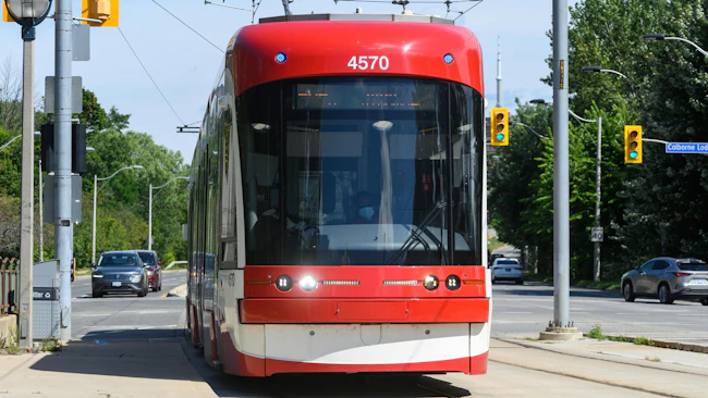 A red and white Toronto Transit Commission Flexity streetcar numbered 4570 travels along a street-level dedicated right-of-way in Toronto, with the CN Tower visible in the distance and traffic signals and trees flanking the tracks.