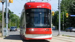 A red and white Toronto Transit Commission Flexity streetcar numbered 4570 travels along a street-level dedicated right-of-way in Toronto, with the CN Tower visible in the distance and traffic signals and trees flanking the tracks.