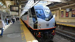 NJ Transit ALP-46 electric locomotive number 4029 in the agency's black, silver, and orange livery sits at a platform at Newark Penn Station, with commuters walking alongside the train and wayfinding signs for PATH, Raymond Boulevard, and the waiting room visible overhead.
