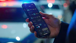 A tight crop of a person's hand holding an iPhone displaying the Cloundrunner App search function. They're standing in front of a blurred police vehicle at night with its lights flashing blue and red.