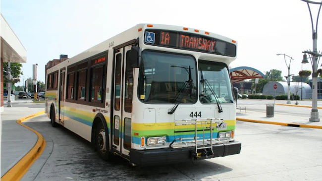A city of Windsor Transit bus number 444 with a white exterior and green, yellow, and blue horizontal striped livery displays '1A Transway' on its destination board while stopped at a transit terminal, with a front-mounted bicycle rack and ADA accessibility symbol visible.