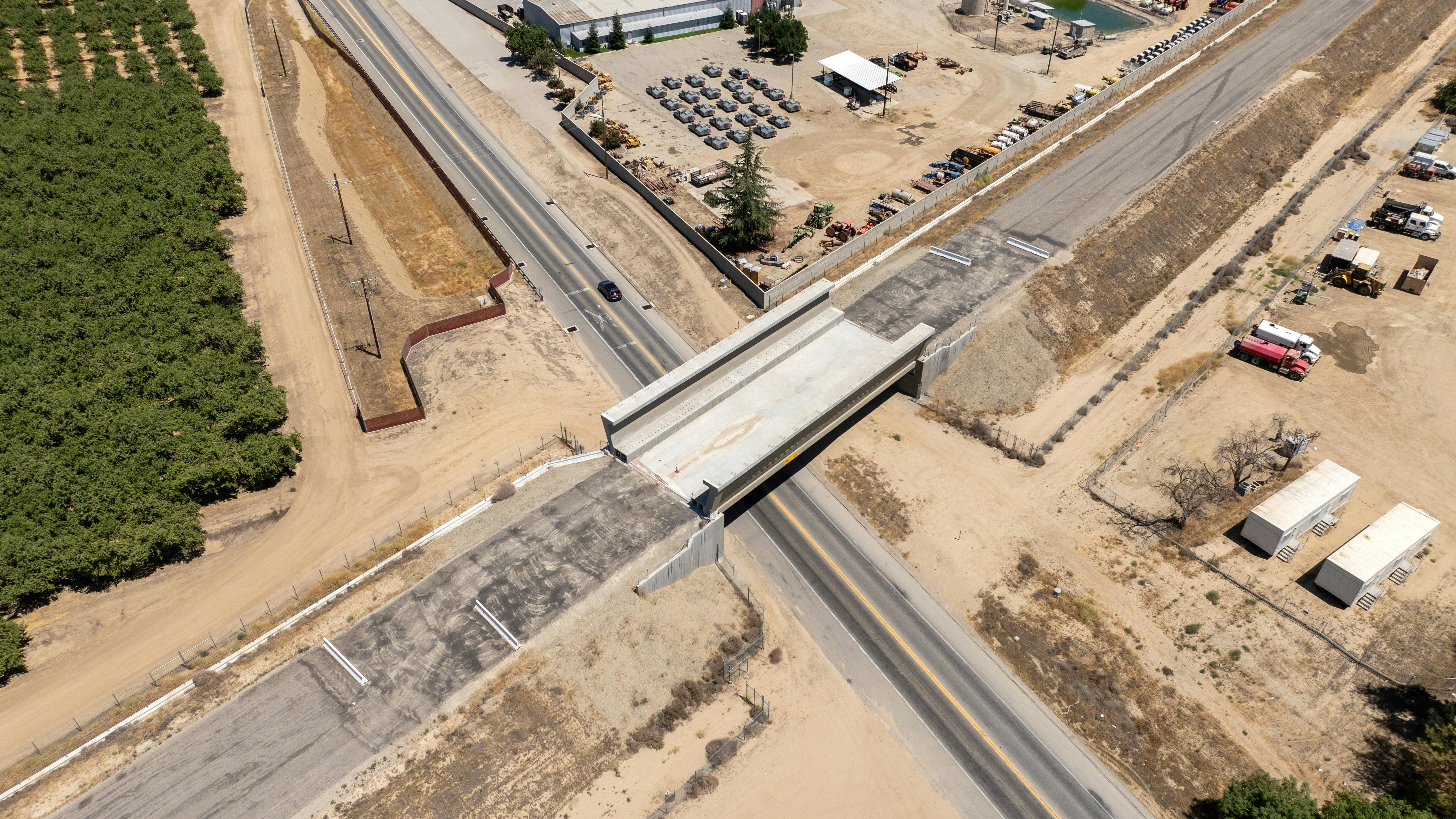 An aerial view of the newly completed Kimberlina Road grade separation viaduct for the California High-Speed Rail project, a concrete structure spanning a two-lane roadway with almond orchards, industrial equipment yards and Central Valley farmland flanking either side.