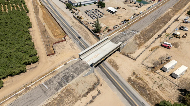 An aerial view of the newly completed Kimberlina Road grade separation viaduct for the California High-Speed Rail project, a concrete structure spanning a two-lane roadway with almond orchards, industrial equipment yards and Central Valley farmland flanking either side.