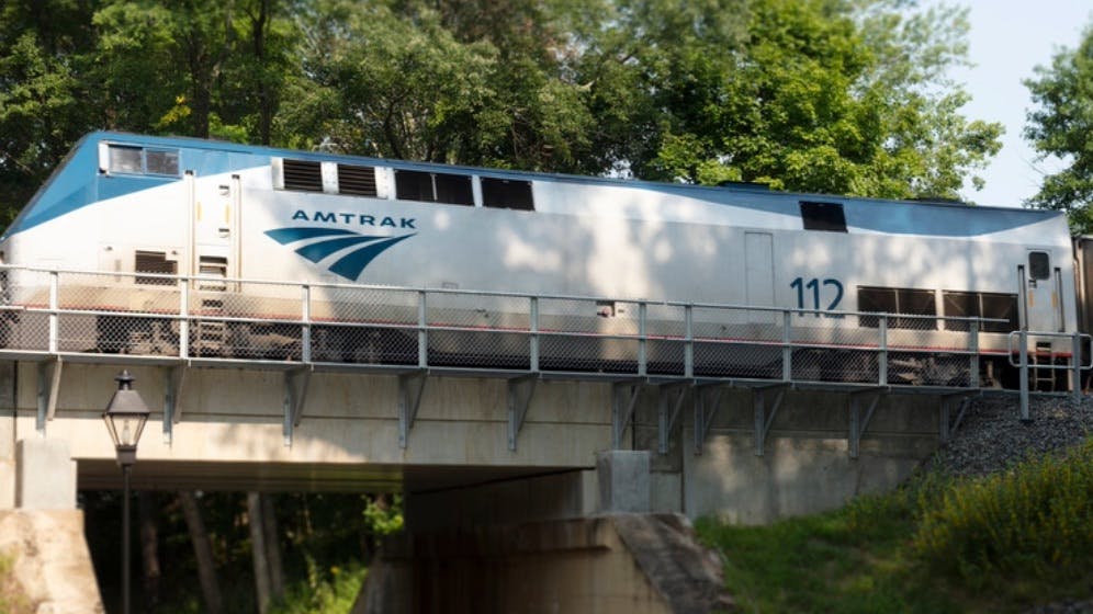 A silver and blue Amtrak GE Genesis P42DC locomotive numbered 112 crosses a small stone and steel bridge over a wooded ravine, with a coach car following behind and green foliage lining the tracks.