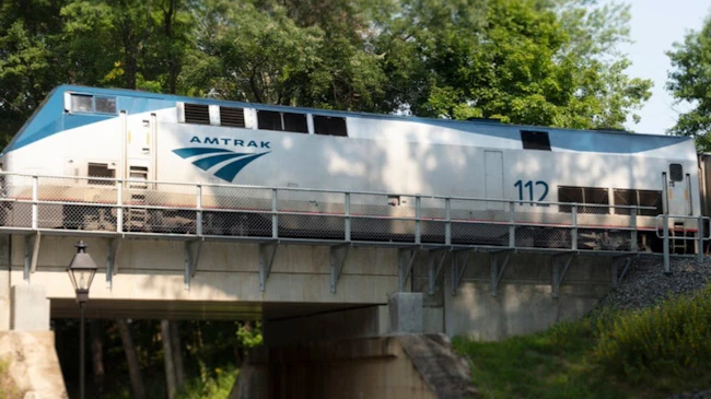 A silver and blue Amtrak GE Genesis P42DC locomotive numbered 112 crosses a small stone and steel bridge over a wooded ravine, with a coach car following behind and green foliage lining the tracks.