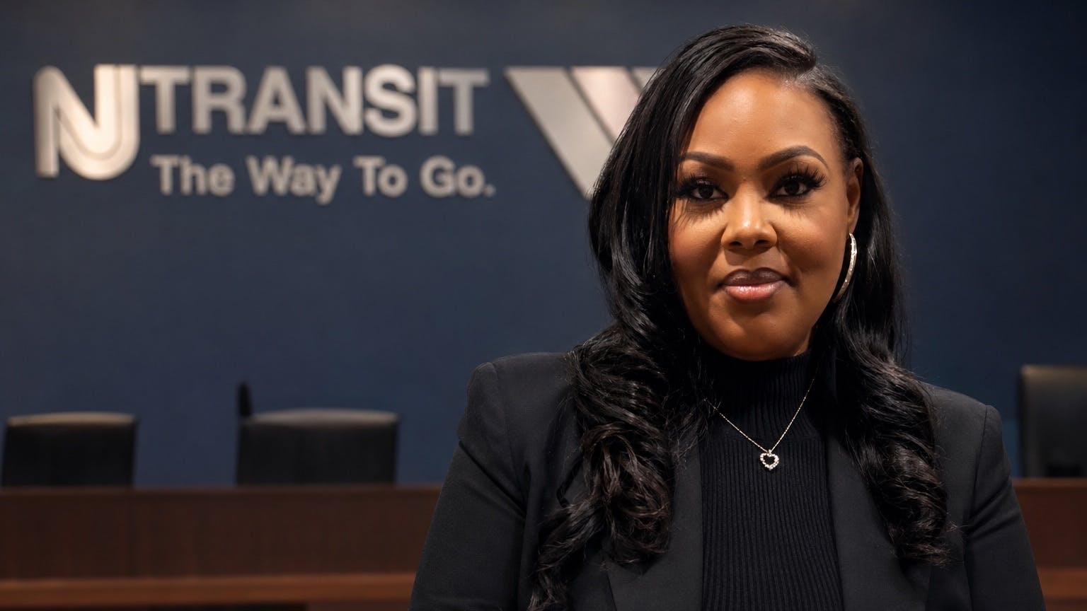 Latashia S. Vauss stands in black professional clothing in front of a blurred New Jersey Transit logo.