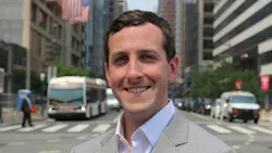 Ryan T. Judge stands in a suit in front of a blurred intersection with a bus in the background.