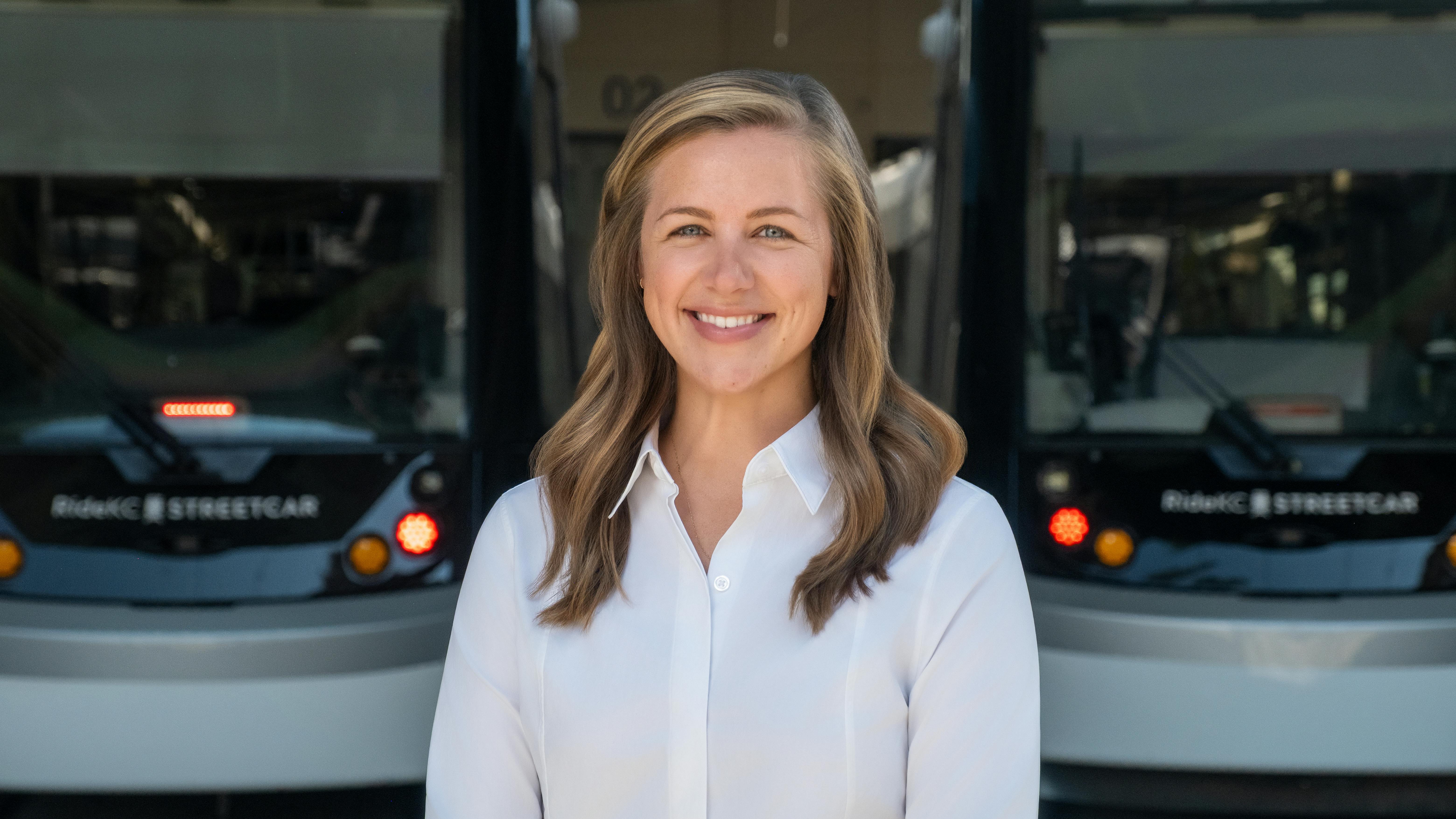 Lauren Krutty, AICP, stands in front of a pair of blurred streetcars. She's smiling and wearing a white blouse.