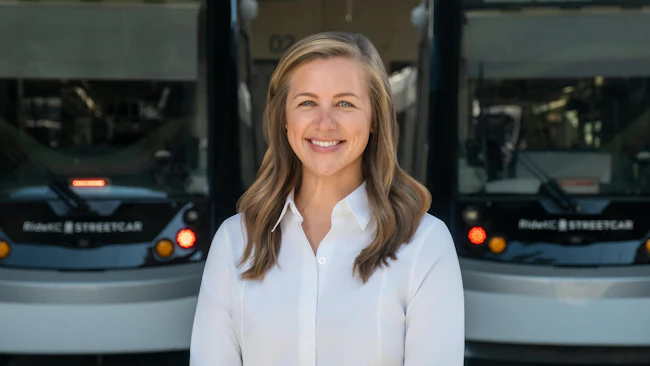 Lauren Krutty, AICP, stands in front of a pair of blurred streetcars. She's smiling and wearing a white blouse.