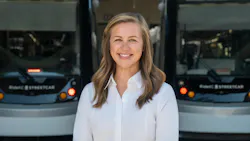 Lauren Krutty, AICP, stands in front of a pair of blurred streetcars. She's smiling and wearing a white blouse.