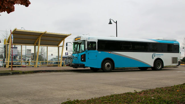 Cherriots Regional bus number 364 in a white and teal livery displays Route 80X on its destination board while pulling away from a yellow-framed bus shelter on an overcast day in the Salem, Oregon area.
