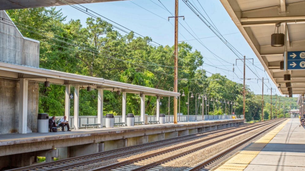 A platform view at BWI Thurgood Marshall Airport Amtrak/MARC station in Maryland, showing two covered island platforms flanking multiple Northeast Corridor tracks, with catenary wire infrastructure overhead, lush green trees in the background, and wayfinding signage directing passengers toward the elevator, overpasses, and tracks toward Washington.