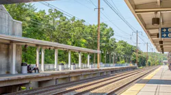 A platform view at BWI Thurgood Marshall Airport Amtrak/MARC station in Maryland, showing two covered island platforms flanking multiple Northeast Corridor tracks, with catenary wire infrastructure overhead, lush green trees in the background, and wayfinding signage directing passengers toward the elevator, overpasses, and tracks toward Washington.