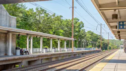A platform view at BWI Thurgood Marshall Airport Amtrak/MARC station in Maryland, showing two covered island platforms flanking multiple Northeast Corridor tracks, with catenary wire infrastructure overhead, lush green trees in the background, and wayfinding signage directing passengers toward the elevator, overpasses, and tracks toward Washington.