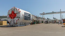 A large white Herrenknecht tunnel boring machine with a red cutterhead is staged at a manufacturing facility, with a sign reading 'Tunnelling for the Yonge North Subway Extension' and logos of Canada, Ontario, Metrolinx and project partners displayed on the machine's side.