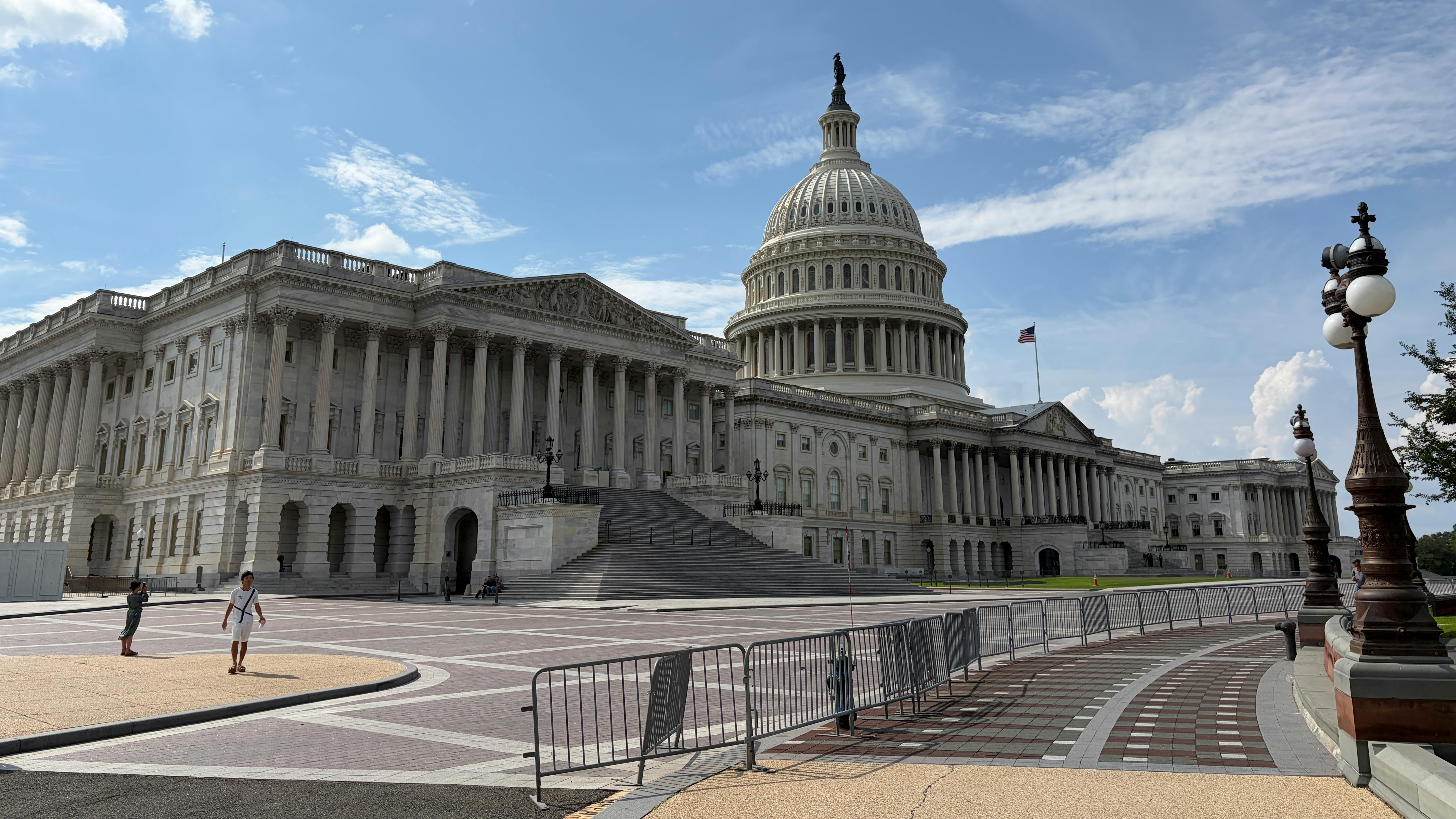 The U.S. Capitol Building's east front, showing the columned Senate wing at left, the central dome topped by the Statue of Freedom and an American flag flying above, with pedestrians walking across the plaza in the foreground.