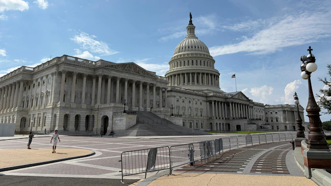The U.S. Capitol Building's east front, showing the columned Senate wing at left, the central dome topped by the Statue of Freedom and an American flag flying above, with pedestrians walking across the plaza in the foreground.