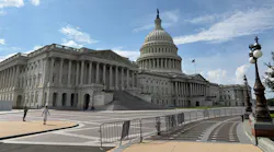 The U.S. Capitol Building's east front, showing the columned Senate wing at left, the central dome topped by the Statue of Freedom and an American flag flying above, with pedestrians walking across the plaza in the foreground.