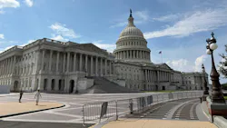 The U.S. Capitol Building's east front, showing the columned Senate wing at left, the central dome topped by the Statue of Freedom and an American flag flying above, with pedestrians walking across the plaza in the foreground.
