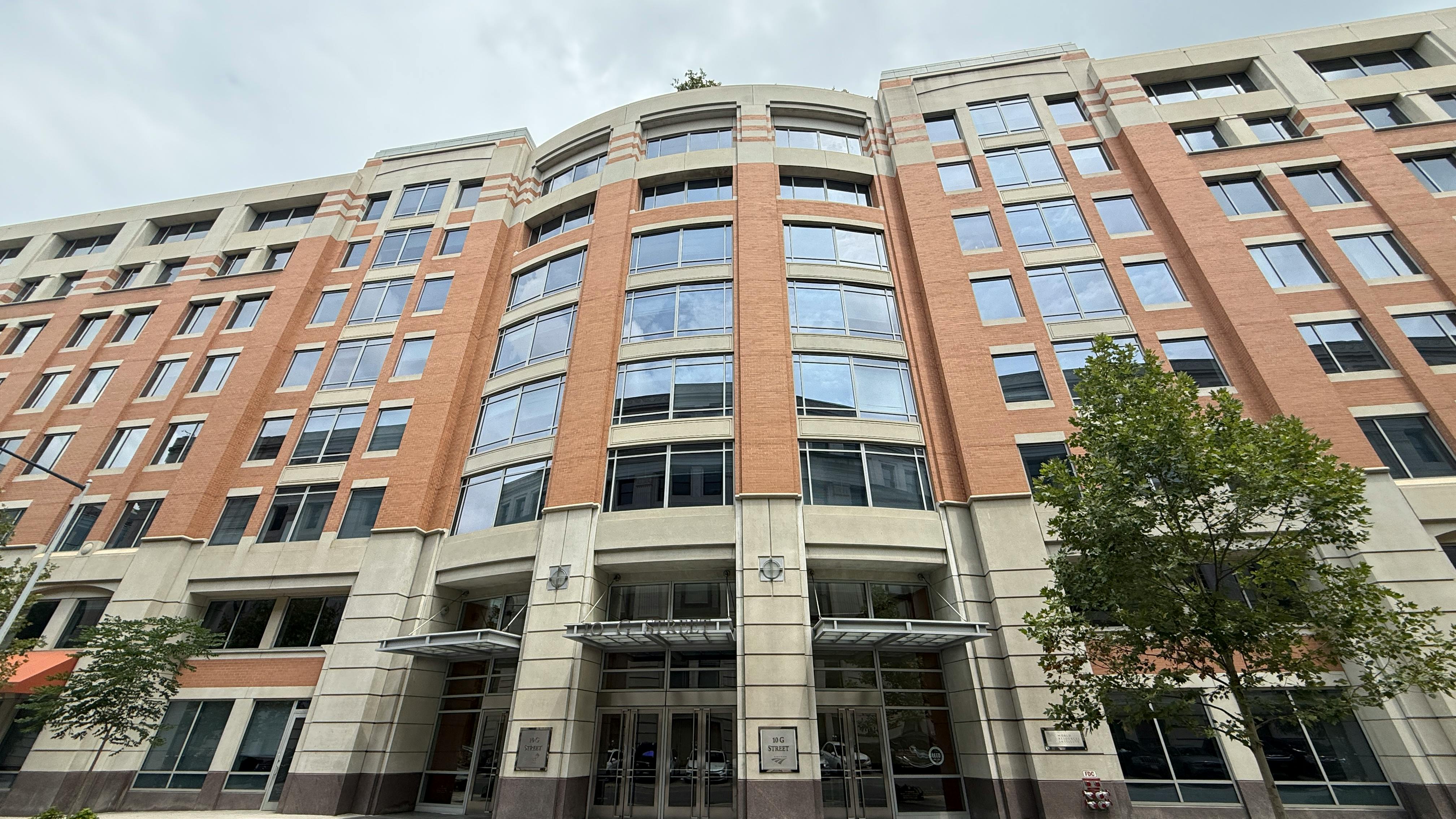 A wide street-level view of the full facade of 10 G Street NE, Amtrak's headquarters in Washington, D.C., a multi-story brick and stone office building with large curved glass windows, a central entrance canopy, and street trees flanking the exterior.