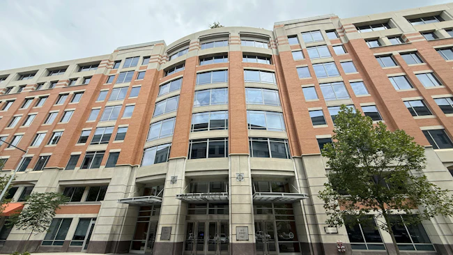 A wide street-level view of the full facade of 10 G Street NE, Amtrak's headquarters in Washington, D.C., a multi-story brick and stone office building with large curved glass windows, a central entrance canopy, and street trees flanking the exterior.