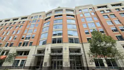 A wide street-level view of the full facade of 10 G Street NE, Amtrak's headquarters in Washington, D.C., a multi-story brick and stone office building with large curved glass windows, a central entrance canopy, and street trees flanking the exterior.