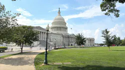The U.S. Capitol Building framed by tree branches overhead, with the central dome topped by the Statue of Freedom and an American flag flying alongside, viewed from across a grassy lawn with a pathway and vintage lamppost in the foreground.