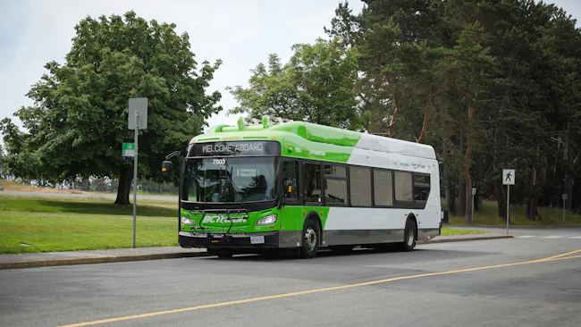 A green and white BC Transit New Flyer battery-electric bus numbered 7503 with 'Welcome Aboard' on its destination sign pulls up to a bus stop, with tall evergreen trees and a grassy area behind.