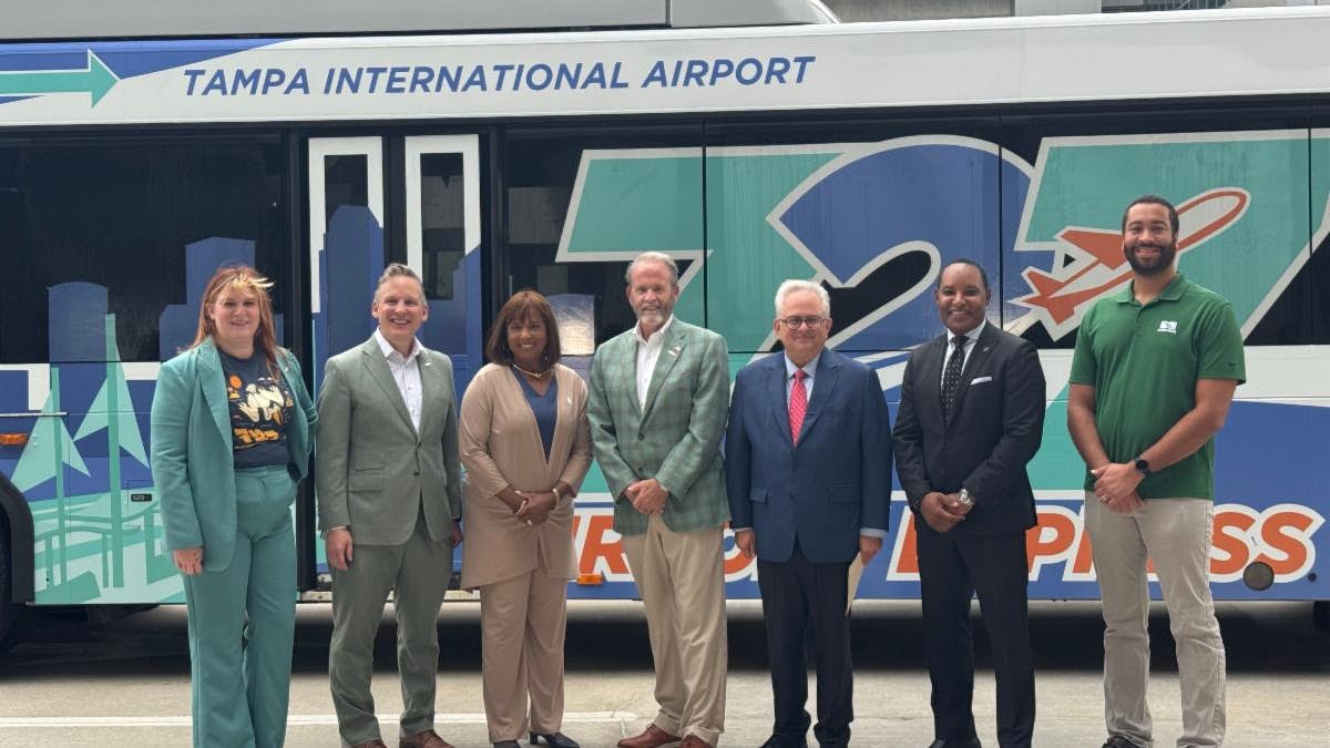 Seven transit and airport officials pose in front of a 727 Express bus bearing 'Tampa International Airport' branding and a teal and blue Tampa skyline wrap, at an announcement event in Tampa, Fla.