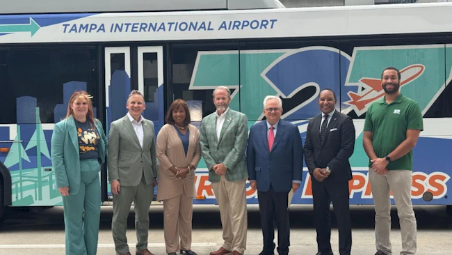 Seven transit and airport officials pose in front of a 727 Express bus bearing 'Tampa International Airport' branding and a teal and blue Tampa skyline wrap, at an announcement event in Tampa, Fla.