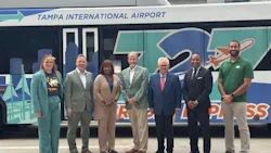 Seven transit and airport officials pose in front of a 727 Express bus bearing 'Tampa International Airport' branding and a teal and blue Tampa skyline wrap, at an announcement event in Tampa, Fla.