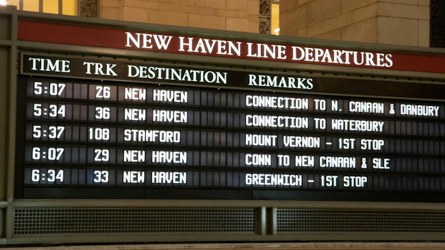 A Metro-North Railroad New Haven Line departures board at Grand Central Terminal displaying upcoming train times, tracks, destinations and connection details, with the terminal's warm-toned stone walls and a chandelier visible above.