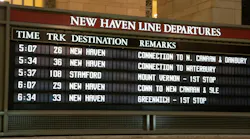 A Metro-North Railroad New Haven Line departures board at Grand Central Terminal displaying upcoming train times, tracks, destinations and connection details, with the terminal's warm-toned stone walls and a chandelier visible above.