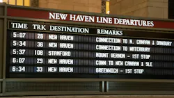 A Metro-North Railroad New Haven Line departures board at Grand Central Terminal displaying upcoming train times, tracks, destinations and connection details, with the terminal's warm-toned stone walls and a chandelier visible above.