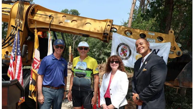 Prince George's County Executive Steuart Pittman, Bike AAA President Jon Korin, DPW Director Karen Henry, and Transportation Officer Sam Snead pose in front of a yellow excavator and Maryland state flag at the ceremonial groundbreaking for a new Transportation Operations Facility.