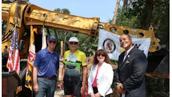 Prince George's County Executive Steuart Pittman, Bike AAA President Jon Korin, DPW Director Karen Henry, and Transportation Officer Sam Snead pose in front of a yellow excavator and Maryland state flag at the ceremonial groundbreaking for a new Transportation Operations Facility.