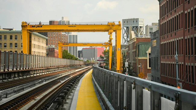 Two large yellow Mi-Jack Travelift gantry cranes span elevated commuter rail tracks along the Park Avenue Viaduct in New York, with construction access walkways lined with yellow safety surfacing running alongside the rails and Harlem brownstones and mid-rise buildings flanking the corridor.