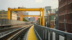 Two large yellow Mi-Jack Travelift gantry cranes span elevated commuter rail tracks along the Park Avenue Viaduct in New York, with construction access walkways lined with yellow safety surfacing running alongside the rails and Harlem brownstones and mid-rise buildings flanking the corridor.
