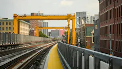 Two large yellow Mi-Jack Travelift gantry cranes span elevated commuter rail tracks along the Park Avenue Viaduct in New York, with construction access walkways lined with yellow safety surfacing running alongside the rails and Harlem brownstones and mid-rise buildings flanking the corridor.