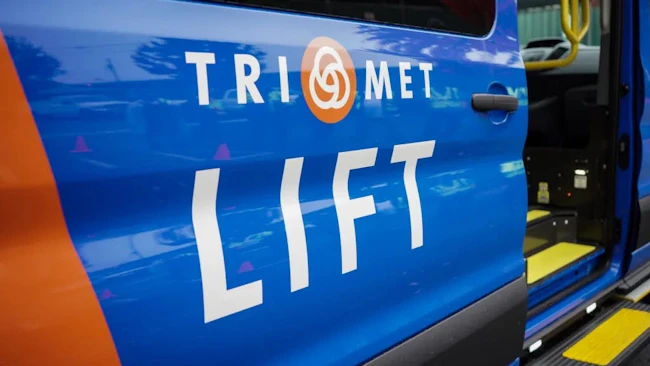 A close-up of a blue and orange TriMet LIFT paratransit vehicle's open side door, with the TriMet logo and 'LIFT' service name displayed prominently and a yellow grab bar and boarding step visible inside.