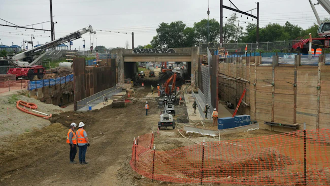 An active rail infrastructure construction site showing a deep excavation corridor beneath an existing bridge structure, with construction workers in orange safety vests and hard hats, cranes, heavy equipment, sheet pile walls, and catenary wire infrastructure visible overhead, with a commuter rail station platform visible in the background to the left.