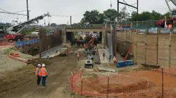An active rail infrastructure construction site showing a deep excavation corridor beneath an existing bridge structure, with construction workers in orange safety vests and hard hats, cranes, heavy equipment, sheet pile walls, and catenary wire infrastructure visible overhead, with a commuter rail station platform visible in the background to the left.