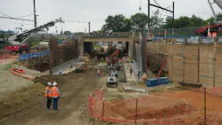 An active rail infrastructure construction site showing a deep excavation corridor beneath an existing bridge structure, with construction workers in orange safety vests and hard hats, cranes, heavy equipment, sheet pile walls, and catenary wire infrastructure visible overhead, with a commuter rail station platform visible in the background to the left.