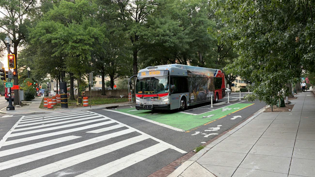A WMATA Metrobus numbered 715 displaying Route D98 to Bethesda on its destination board travels through a green-painted bike lane at a tree-lined urban intersection in Washington, D.C., with construction barrels visible on the left and ADA-compliant curb ramps in the foreground.