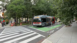 A WMATA Metrobus numbered 715 displaying Route D98 to Bethesda on its destination board travels through a green-painted bike lane at a tree-lined urban intersection in Washington, D.C., with construction barrels visible on the left and ADA-compliant curb ramps in the foreground.