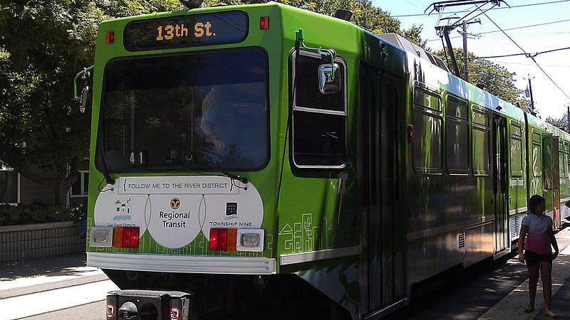 A green Sacramento Regional Transit Green Line light rail vehicle numbered 41 with a '13th St.' destination sign and a 'Follow me to the River District' promotional wrap sits at a station platform, with a rider standing to the right and overhead catenary wires above.