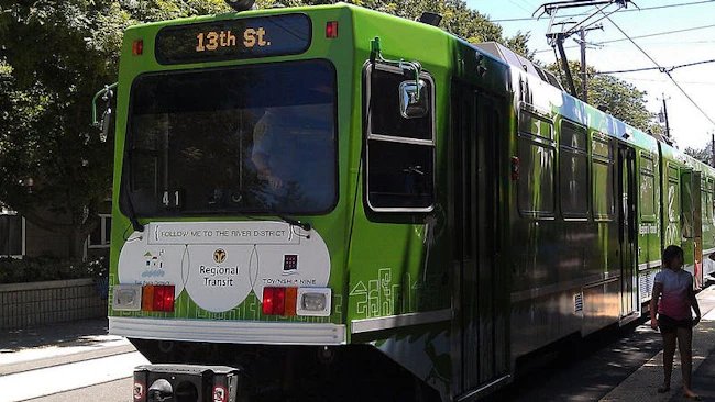 A green Sacramento Regional Transit Green Line light rail vehicle numbered 41 with a '13th St.' destination sign and a 'Follow me to the River District' promotional wrap sits at a station platform, with a rider standing to the right and overhead catenary wires above.