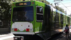 A green Sacramento Regional Transit Green Line light rail vehicle numbered 41 with a '13th St.' destination sign and a 'Follow me to the River District' promotional wrap sits at a station platform, with a rider standing to the right and overhead catenary wires above.