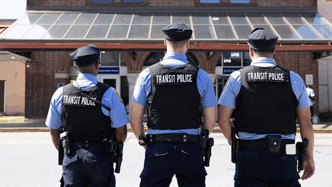 Three SEPTA Transit Police officers stand with their backs to the camera in front of a brick transportation center, wearing blue uniforms, ballistic vests bearing 'Transit Police' identification and duty belts with holstered sidearms.