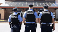Three SEPTA Transit Police officers stand with their backs to the camera in front of a brick transportation center, wearing blue uniforms, ballistic vests bearing 'Transit Police' identification and duty belts with holstered sidearms.